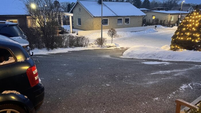 Snötäckt uppfart med bilar, hus och en julgran med ljus i skymningen.