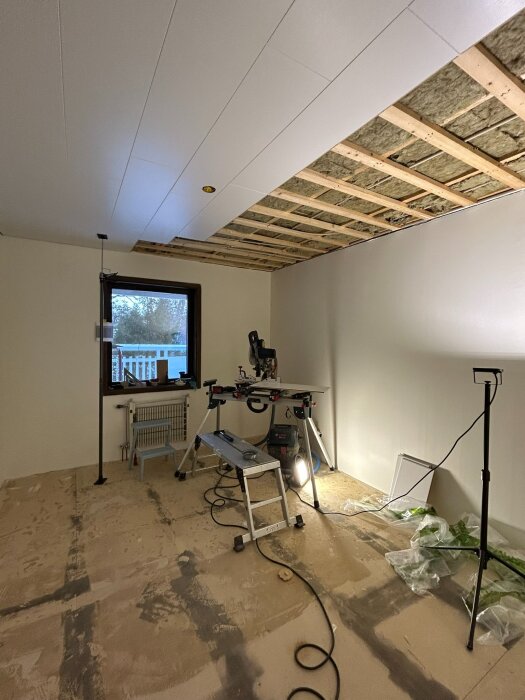 Rum under renovering med halvfärdigt innertak och verktyg på arbetsbord; golvet avskalat redo för renovering, stege och belysning närvarande.
