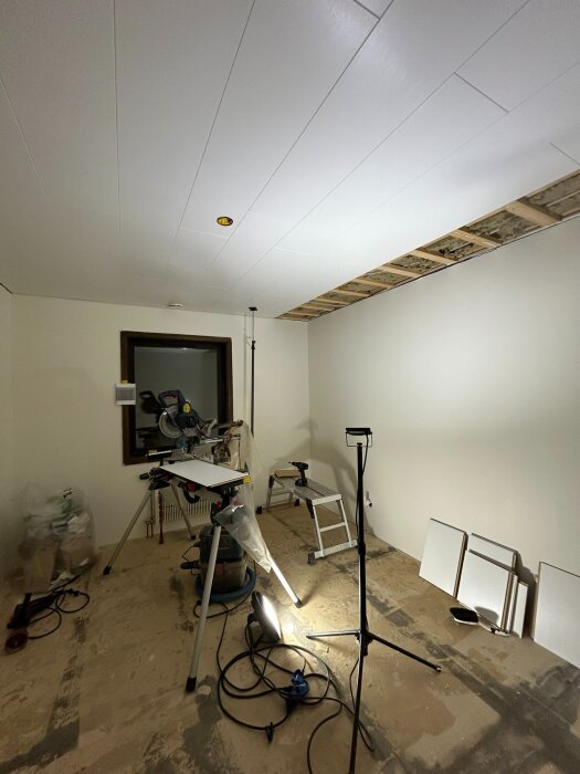 Rum under renovering med nytt tak, sågbänk och verktyg på golvet. Väggar och golv ofärdiga med påbörjat arbete.