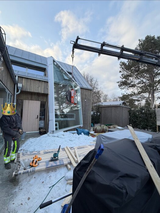 Kran lyfter stort glasparti på plats vid tillbyggnad, omgiven av trävirke och byggnadsmaterial på snöig mark. Person i skyddskläder tittar på.