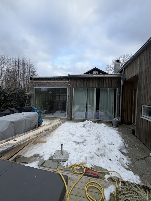 Tillbyggnad under snöig vinterdag med ett stort skjutparti installerat. Byggmaterial ligger på marken och trädgårdsredskap syns i förgrunden.