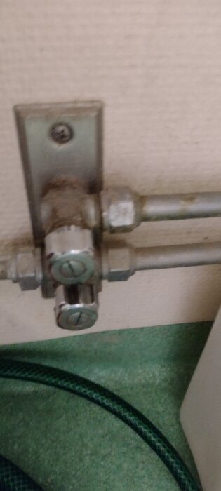 Väggmonterad rörkoppling med två metallventiler, synlig mot en vit vägg och grön slang i förgrunden.