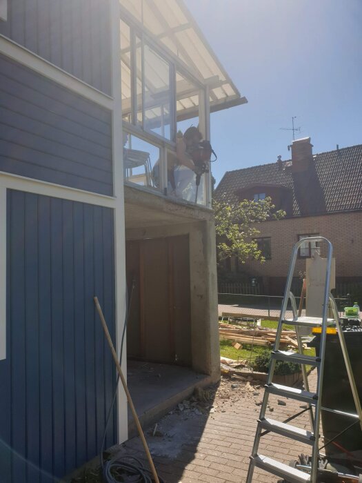 En person arbetar med glasfasaden på ett blått hus, byggnadsställning på marken och verktyg i bakgrunden.