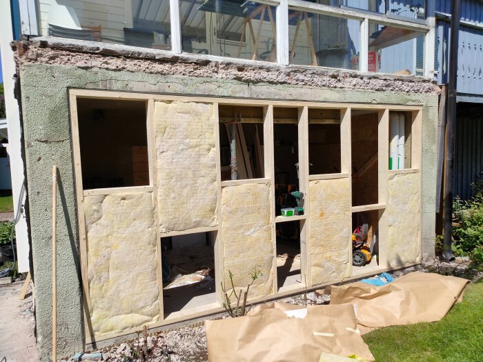 Byggnad under renovering med frilagd stomme och isolering på en vägg, glasfönster ovanför och diverse byggmaterial framför.