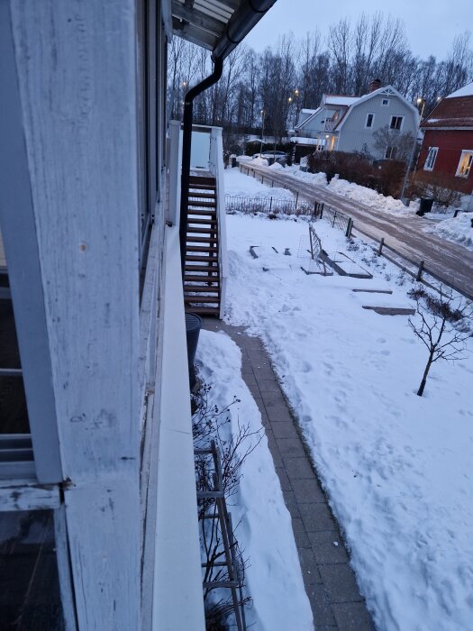 Husfasad i vintermiljö med fönster och stuprör. Snötäckt gångväg och trädgård syns bredvid huset, samt snötäckta träd och vägar längre bort.