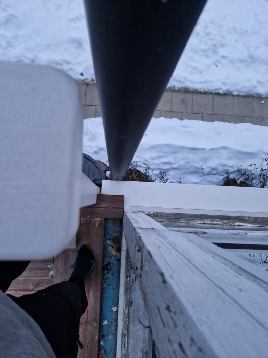 Person står på balkongkant, med strumpor, nära en stång. Snötäckt mark och marksten syns nedanför.