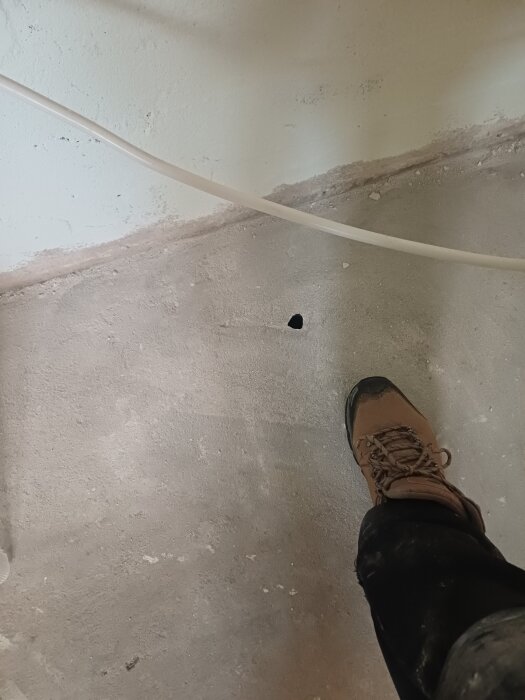 Brun stövel vid ett litet hål i ett betonggolv, ljus vägg och en vit kabel syns i bakgrunden.