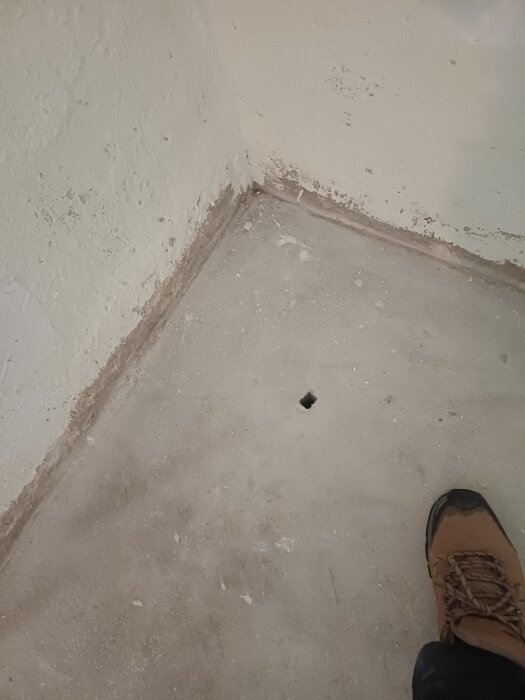 Betonggolv med ett hål i ett rumshörn, ytan är omgiven av vita murade väggar. En brun stövel är synlig i nederkanten av bilden.