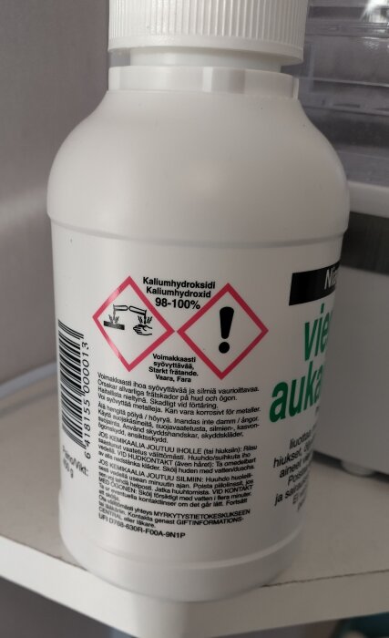 Plastflaska etiketterad "Kaliumhydroxid 98-100%" med varningssymboler för frätande ämnen och faroangivelse på flera språk.