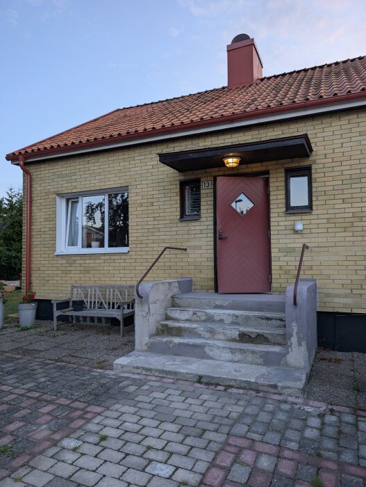Entré med sliten trappa och tegelväggar, röd dörr och tak, samt en träbänk bredvid.
