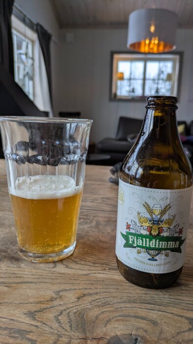 Ölflaska märkt "Fjälldimma" och glas på träbord i stuga.