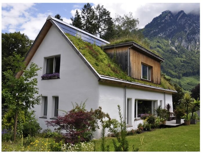 Ett modernt hus med grönt tak och solpaneler, omgivet av grönska och berg i bakgrunden.