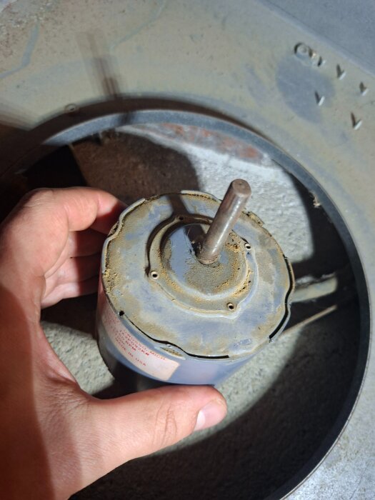 En hand håller en dammig och sliten elektrisk motor med en axel, omgiven av ett metallhölje i en verkstadsmiljö.
