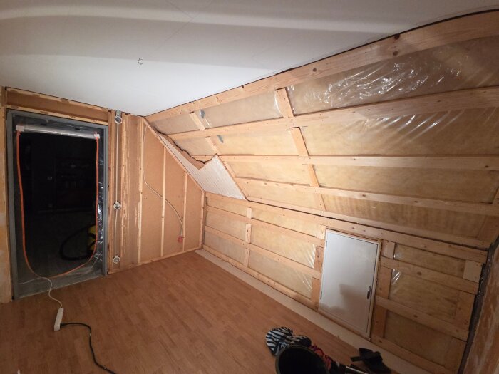 Ett rum under renovering där masonit är borttagen från ena väggen och synliga kablar finns. Голv av träimitation och en liten dörr syns.