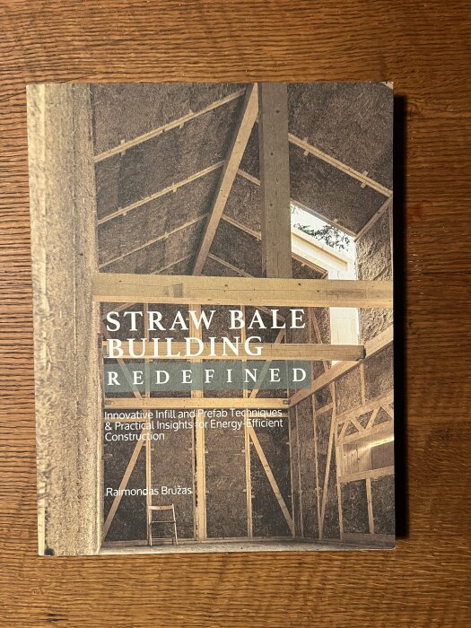 Boken "Straw Bale Building Redefined" ligger på ett träbord, med detaljer om hållbara byggtekniker och hög energieffektivitet.