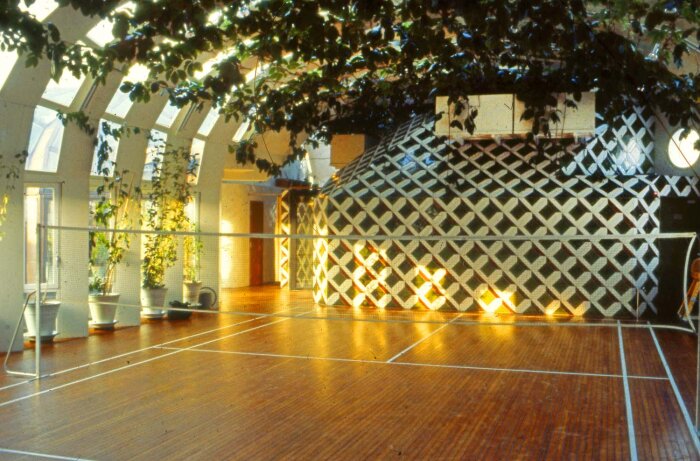 Inomhus tennisbana med glastak och växter, samt ett mönstrat, välvt utrymme i bakgrunden, designat av arkitekt Peter Celsing.