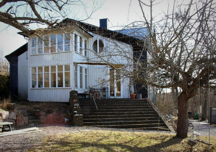 Peter Celsings Villa Klockberga, vit och svart fasad, stora fönster, trappa med krukor, trädgård med träd utan löv.