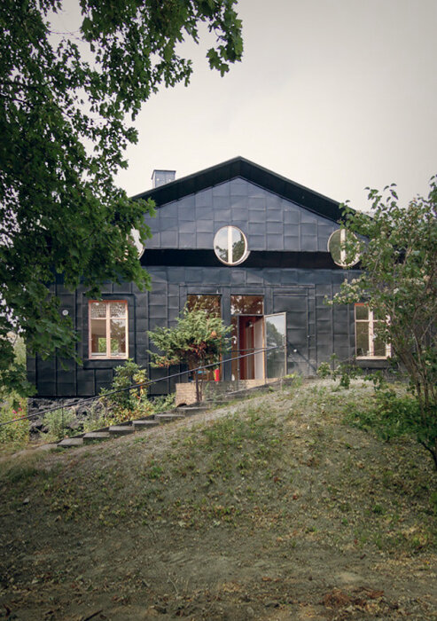Svart hus med gröna växter vid entrén och runda fönster, omgivet av träd på en sluttning.