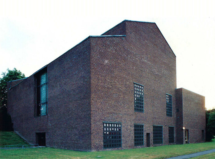 Rödtegelskyrka med modern design och geometriska fönster, omgivet av gräs och träd.