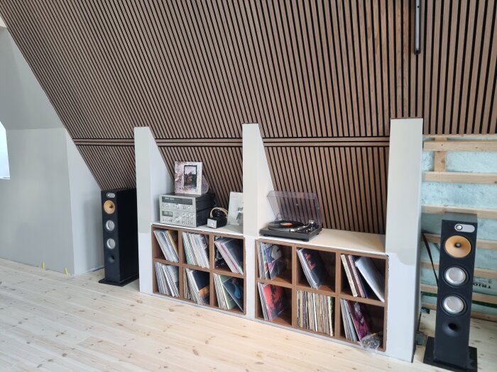 Platsbyggd hylla med vinylspelare och förstärkare, inramad av högtalare, mot en vägg med träpaneler.