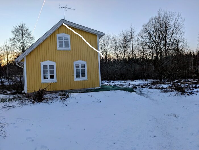 Gul byggnad med asymmetriskt tak, höger sida högre. Snötäckt mark och vinterträd i bakgrunden, en antenn syns på taket.