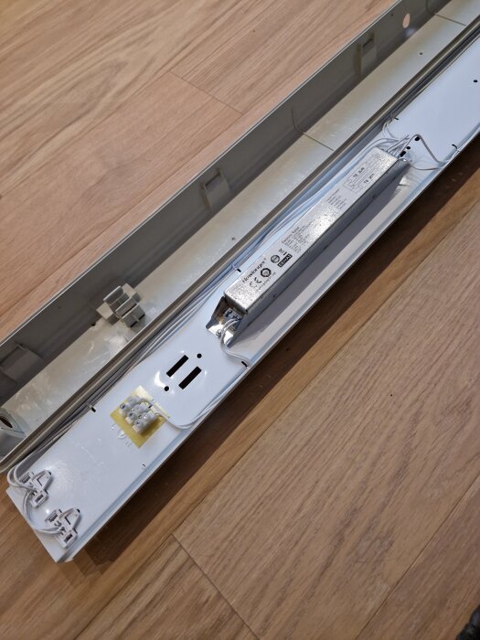 Öppen armaturenhet på golvet med exponerade kablar och drivdon, redo för installation av LED-lysrör i ett garage.