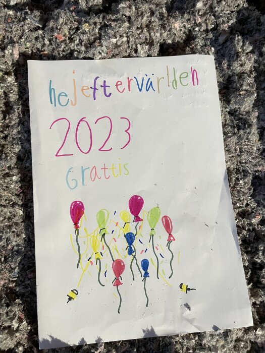 Handskrivet brev med texten "hej eftervärlden 2023, grattis" och ritade ballonger i olika färger på en matta.