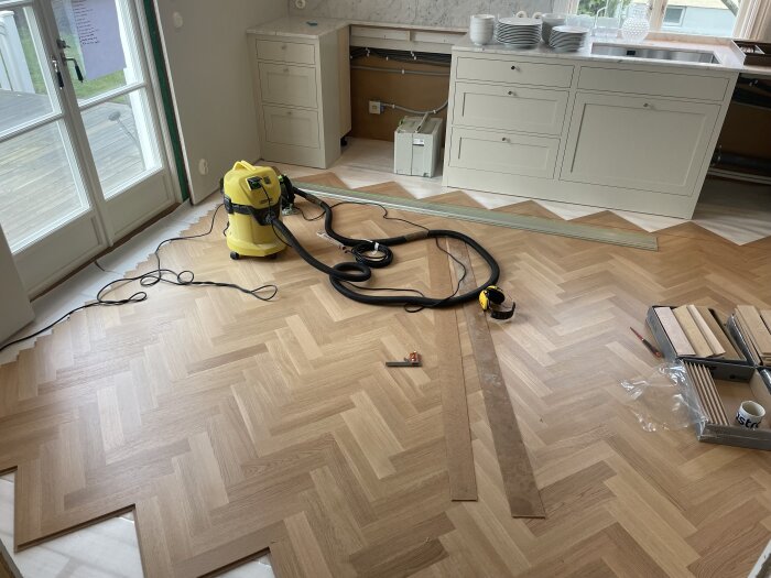 Köksrenovering pågår med nylagd fiskbensparkett, verktyg och en dammsugare på golvet, samt köksinredning i bakgrunden.