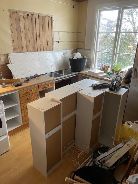 Gamla köket under renovering med borttagen diskbänk och öppnad vägg. Verktyg och material ligger på bänkskivan, med utsikt genom fönstret.