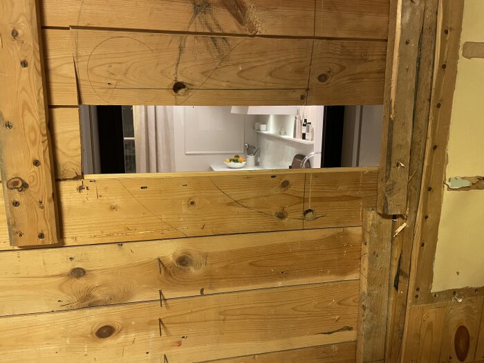 Öppen vägg av träpanel. Utsikt genom hålet visar ett kök med diskbänk, fruktfat och ljus inredning.