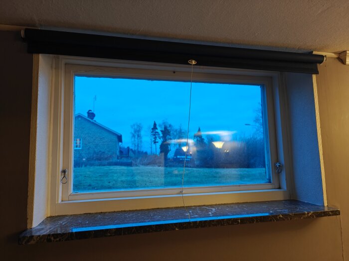 Ett fönster med en mörk rullgardin och marmorfönsterbräda, utsikt mot en trädgård och ett hus i blått skymningsljus.