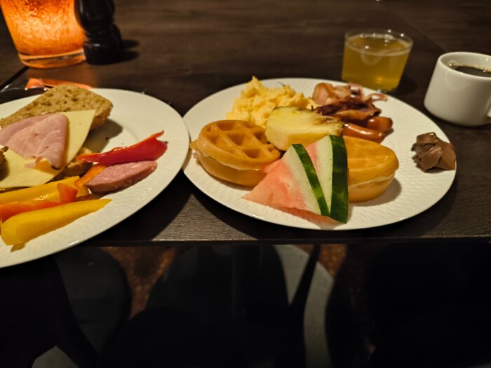 Frukostbuffé med bröd, ost, skinka, paprika, våfflor, vattenmelon, ananas, äggröra, bacon, korv och drycker på ett bord.