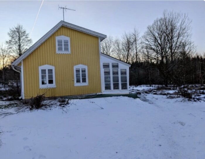 Gult hus med glasveranda och olika takvinklar, omgiven av snöklädd mark och träd i bakgrunden.