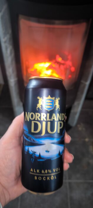 Hand som håller en ölburk framför en brinnande eldstad. Burken har texten "Norrlands Djup Bocköl" med en illustration av en vintersjö.