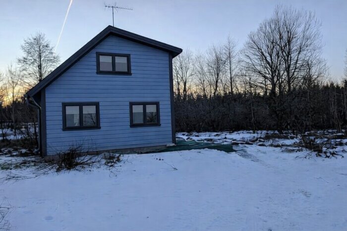 Blått tvåvåningshus med klassisk design, snötäckta omgivningar och skog i bakgrunden, fotograferat vid solnedgång.