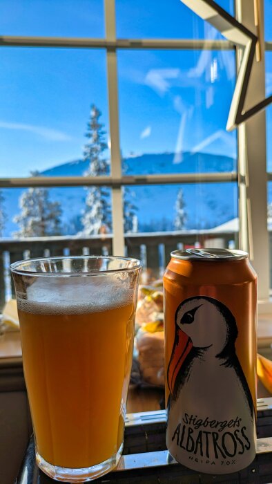 Ett glas och en burk Stigbergets Albatross öl vid ett fönster med utsikt över snötäckta träd och berg i solsken.
