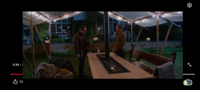 Två personer samtalar vid ett bord under ett tält på en upplyst gård, med en eldstad installerad i bordet.