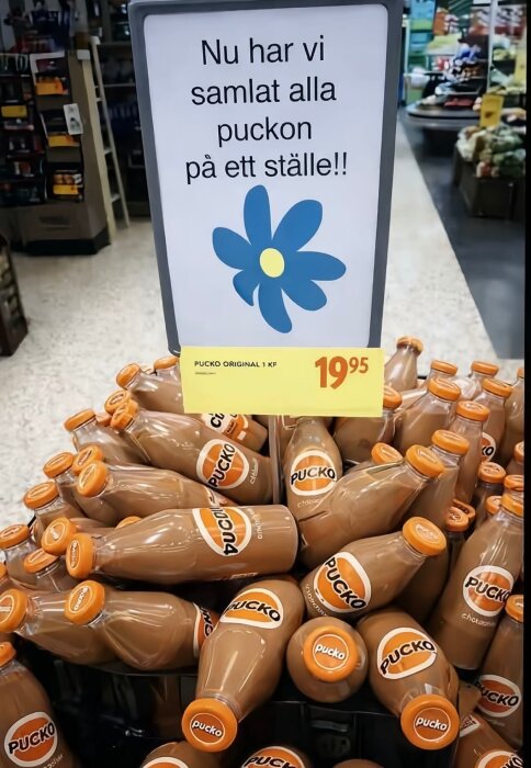 Flaskor med chokladdrycken Pucko samlade i en butik, skylt med humoristisk text och en blå blomma ovanför.