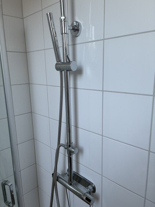 Dusch med kromad duschstång och vitkaklade väggar. Duschhandtag och termostatblandare syns.