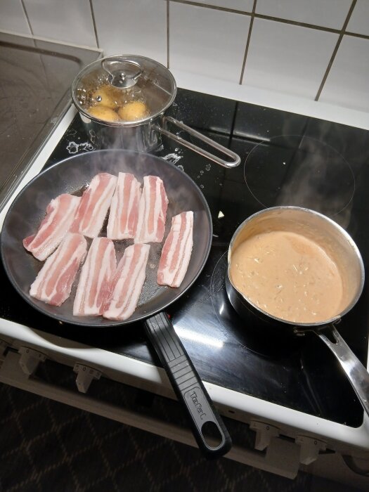 Stekpanna med rått fläsk, kastrull med löksås och kokande potatis på spis.