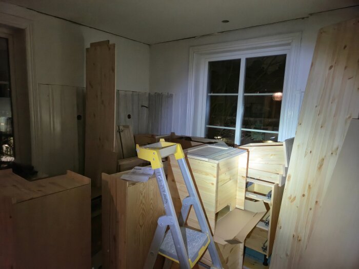 Rum fyllt med trälådor och byggmaterial under köksmontering, med en stege vid ett fönster på kvällstid.