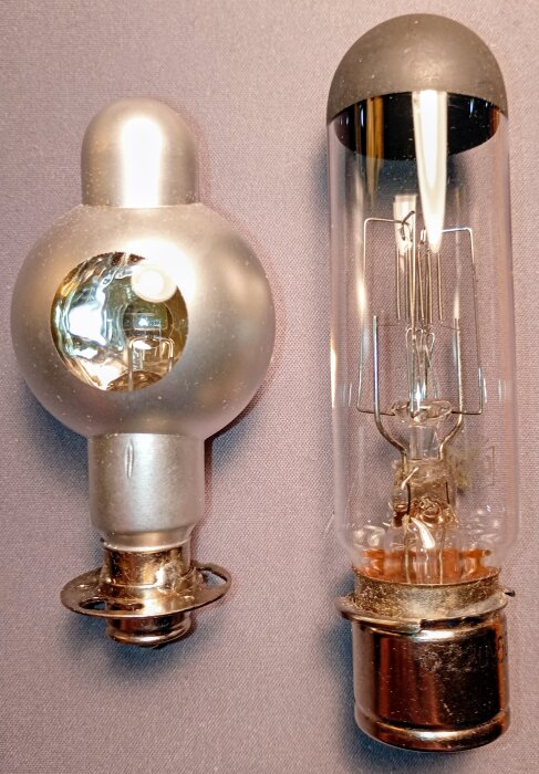 Två gamla radiorör, en grå med sfärisk form och en transparent cylindrisk, ligger bredvid varandra på en grå yta.