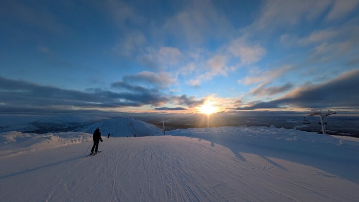 En skidåkare på en snötäckt bergstopp med solnedgång i bakgrunden. Himlen är blå med få moln och en stolslift syns till höger.