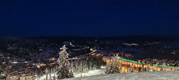 Snötäckt skidbacke med vinterlandskap i bakgrunden och upplyst skidby, tagen på natten med klar mörk himmel.