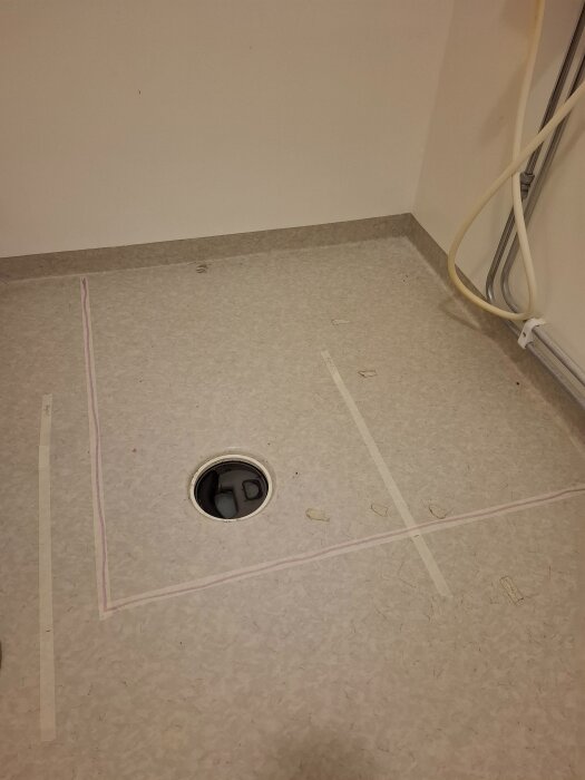Badrumsplanering med tejpmarkerade duschväggar och golvreglar, synlig golvbrunn i mitten, beige golv och vägg vid renovering.
