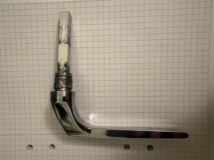 Kromat dörrhandtag av metall, borttaget från balkongdörr, ligger mot rutat papper.