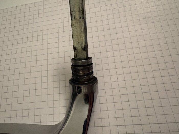 Balkongdörrshandtag av metall, utan låsring, demonterad mot rutigt papper.