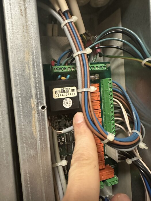 Närbild på styrkort inuti ventilationsaggregat, där en person pekar på komponenter och kablar, med synliga terminaler och ledningar.