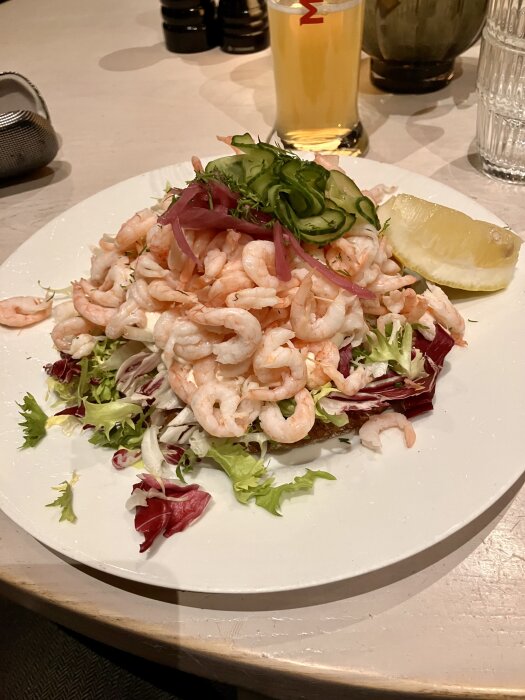 Räksmörgås med sallad, skivad gurka, rödlök och citron på en tallrik, med ett glas öl i bakgrunden.