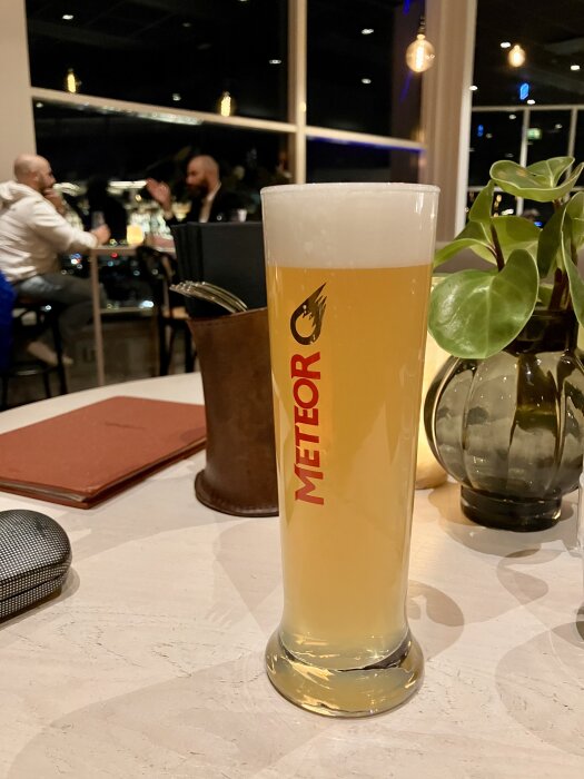 Högt glas med ljus öl märkt "Meteor" på ett cafébord, omgiven av en växt, en plånbok och bakgrund med två personer i samtal.
